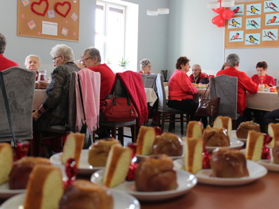 Zdjęcie przedstawia salę główną Domu Dziennego Pobytu Senior+ w Sompolnie. Widać na nim seniorów siedzących przy stole oraz talerze ze słodkim poczęstunkiem.