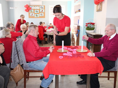 Na zdjęciu dwóch seniorów siedzących przy stole naprzeciwko siebie. Pomiędzy nimi rehabilitantka objaśniająca konkurencję zręcznościową. W tle seniorzy siedzący przy stole.