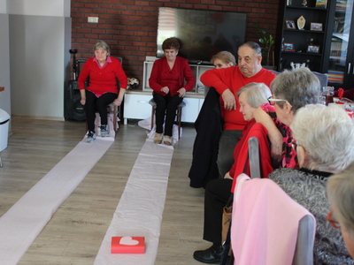 Zdjęcie przedstawia dwie seniorki biorące udział w konkurencji sprawnościowej, polegającej na przysunięciu nogami długiego paska papieru z książką na końcu, w jak najkrótszym czasie.