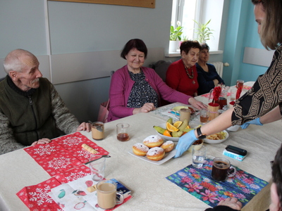 5) Zdjęcie przedstawia seniorów siedzących przy stole i opiekunkę roznoszącą pączki i chruściki.