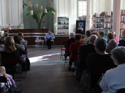 3) Na zdjęciu pisarka siedząca na krześle przy stoliku kawowym  z książką w dłoniach. Po obydwu jej stronach publiczność.