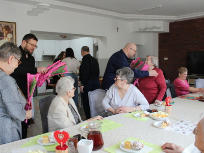 1) Zdjęcie wykonane w sali głównej domu dziennego pobytu senior+ w Sompolnie. Przedstawia mężczyzn wręczających kwiaty seniorkom.