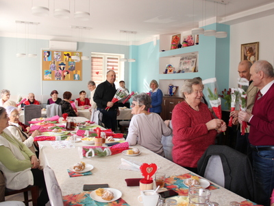 2) Na fotografii kobiety siedzące przy stole oraz mężczyźni składający im życzenia i wręczający kwiaty.