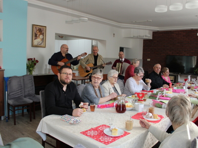 4) Fotografia przedstawia dwóch mężczyzn grających na gitarach i jednego na akordeonie. Widać również uczestników spotkania siedzących przy stole.