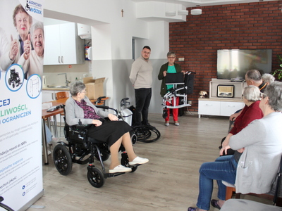 Zdjęcie przedstawia jedną z uczestniczek prezentującą lekkość złożonego wózka, konsultanta medycznego oraz seniorkę siedzącą na wózku.