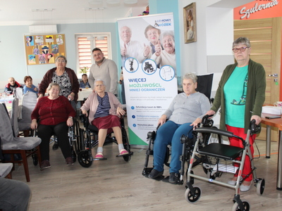 Zdjęcie prezentuje uczestniczki ddp siedzące na wózkach inwalidzkich, panią z balkonikiem oraz konsultanta medycznego zebranych wokół banneru – darmowy wózek.