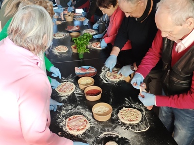 Zdjęcie przedstawia grupę seniorów przy stole wykonujących samodzielnie pizze z dostępnych składkników