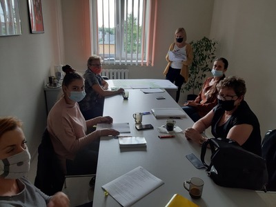 Na zdjęciu znajduje się 6 kobiet. Pięć z nich siedzi przy stole, na którym leżą dokumenty, telefony komórkowe i staja szklanki. Jedna Kobieta stoi z dokumentami w ręku. Wszyscy mają na twarzach maseczki.