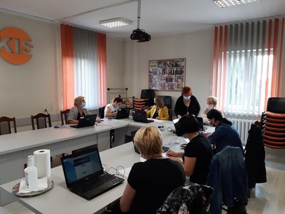 Na zdjęciu znajduje się osiem kobiet siedzących przy stole. Przed każdą stoi otwarty laptop. Jedna kobieta pochyla się przy jednym z laptopów. Wszyscy maja na twarzach maseczki.