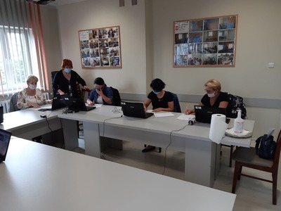 Na zdjęciu znajduje się pięć kobiet siedzących przy stole. Przed każdą stoi otwarty laptop. Jedna kobieta pochyla się przy jednym z laptopów. Wszyscy maja na twarzach maseczki.