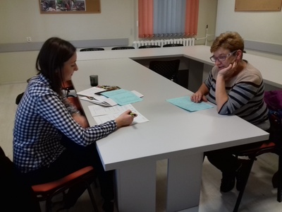 Na zdjęciu znajdują się dwie kobiety. Siedzą przy stole na przeciw siebie. Na stole leżą dokumenty.