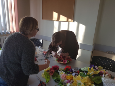 Na zdjęciu znajdują się dwie kobiety stojące przy stole. Na stole leżą sztuczne kwiaty i liście. Kobiety robią stroik.