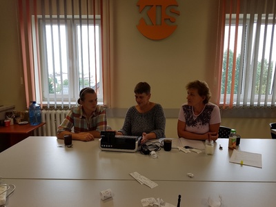 Na zdjęciu znajdują się trzy osoby siedzące przy stole. Przed nimi stoi kasa fiskalna i leżą paragony.