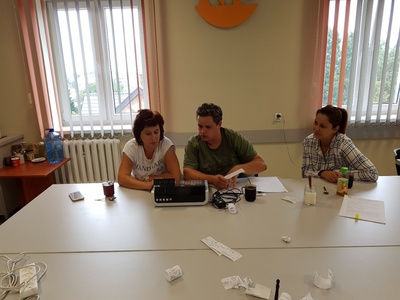 Na zdjęciu znajdują się trzy osoby siedzące przy stole. Przed nimi stoi kasa fiskalna i leżą paragony.