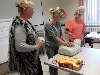 Na zdjęciu znajdują się trzy kobiety. Na stole leży tułów manekina. Jedna z kobiet uczy pozostałe techniki masażu serca.