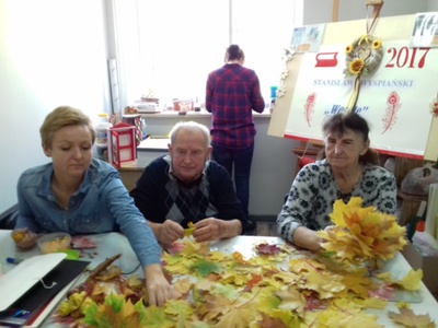 Na zdjęciu znajdują się cztery osoby. Trzy osoby siedzą przy stole, na którym leży dużo suszonych liści. Jedna osoba stoi przy oknie tyłem do pozostałych.