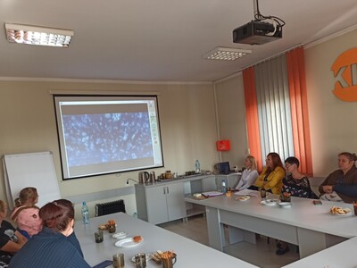 Na zdjęciu znajduje się siedem osób siedzących przy dwóch stołach. Wszyscy patrzą na ekran z prezentacja slajdów.
