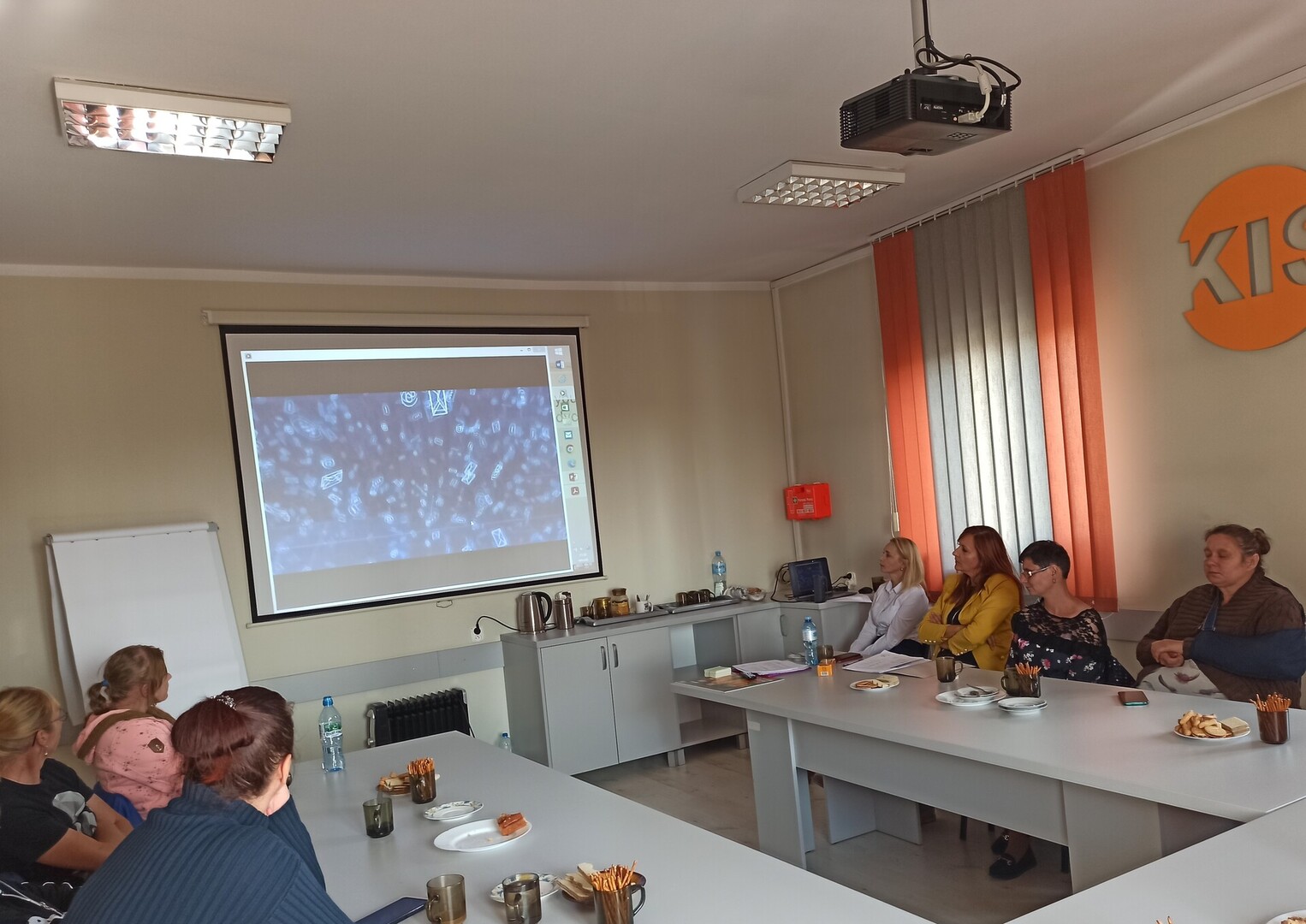 Na zdjęciu znajduje się siedem osób siedzących przy dwóch stołach. Wszyscy patrzą na ekran z prezentacja slajdów. 
