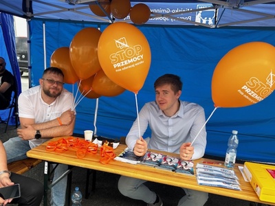 3)	Na fotografii dwaj mężczyźni - pracownicy MGOPS siedzący pod namiotem ośrodka trzymający żółte balony z napisem „Stop przemocy!”. Przed nimi stoisko tematyczne.