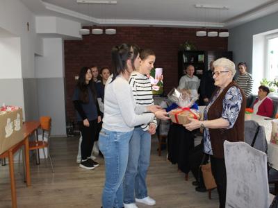 Na fotografii jedna z seniorek przejmuje kartonik z ciastkami. W tle seniorzy i wolontariusze.