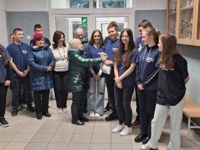 2)	Zdjęcie grupowe na którym jedna z seniorek wręcza wolontariuszom worek z cukierkami. 
