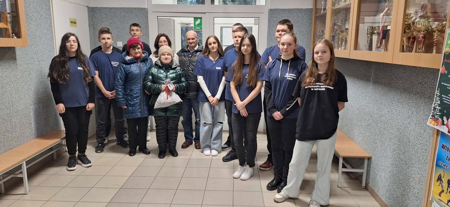 1)	Zdjęcie wykonane na korytarzu szkoły w Mąkolnie. Przedstawia wolontariuszy i seniorów na zdjęciu grupowym.