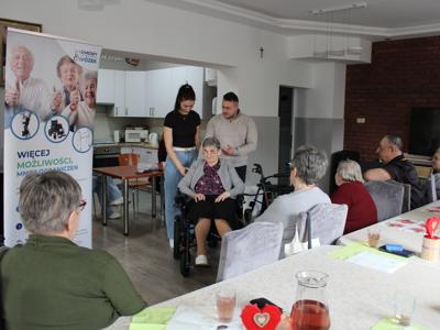 Na zdjęciu fizjoterapeutka i konsultant medyczny prezentują wózek elektryczny na którym siedzi jedna z uczestniczek. Po lewej stronie baner – darmowy wózek oraz seniorzy siedzący przy stole.