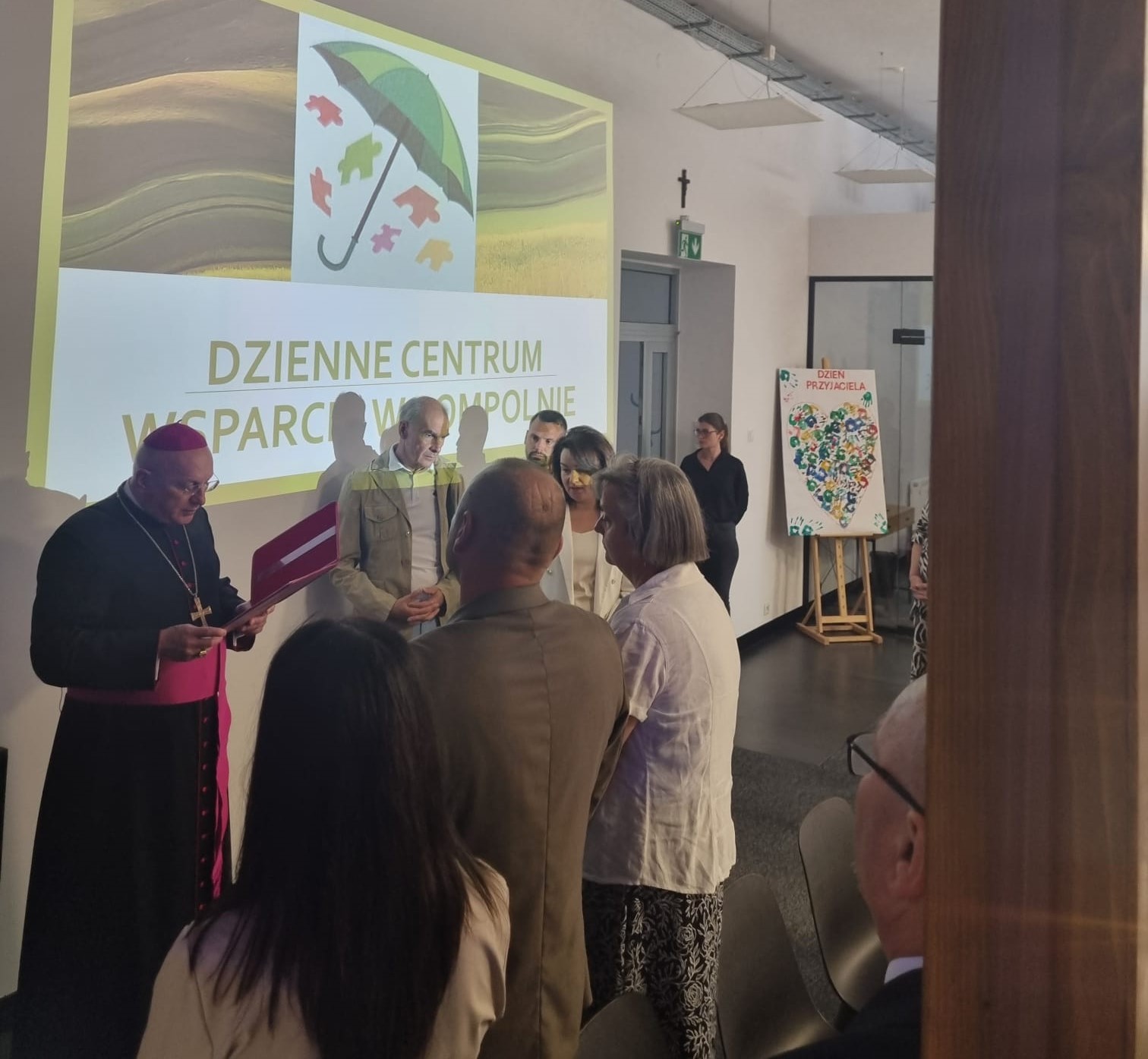 Zdjęcie wykonane w Dziennym Centrum Wsparcia. Przedstawia biskupa oraz przedstawicieli gminy stojących na tle logo placówki.