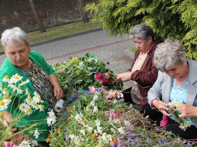 Zdjęcie wykonane na parkingu przy M-GOPS w Sompolnie przedstawia trzy seniorki siedzące przy stole podczas wyplatania wianków