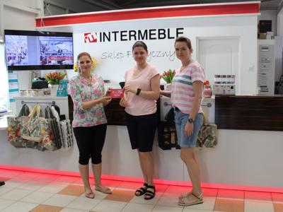 Zdjęcie wykonane wewnątrz sklepu INTERMEBLE. Przedstawia trzy kobiety: dwie z nich wręczają jednej voucher do wykorzystania w sklepie. Za nimi pulpit i logo sklepu.