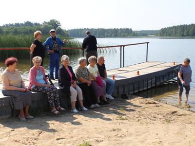 Na zdjęciu seniorzy siedzący na pomoście nad jeziorem lubstowskim. Jedna z seniorek spaceruje na brzegu jeziora podpierając się kulą.