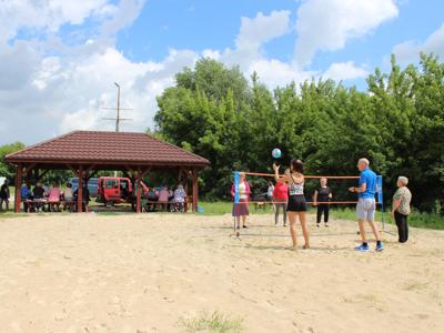 Fotografia przedstawia seniorów grających w piłkę siatkową na plaży oraz altanę grillową pod którą siedzą seniorzy.