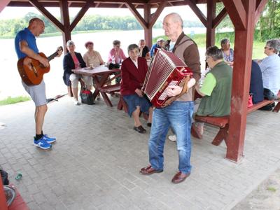 Zdjęcie przedstawia mężczyzn grających na akordeonie i gitarze. W tle seniorzy siedzący na ławkach przy stołach pod altaną grillową.