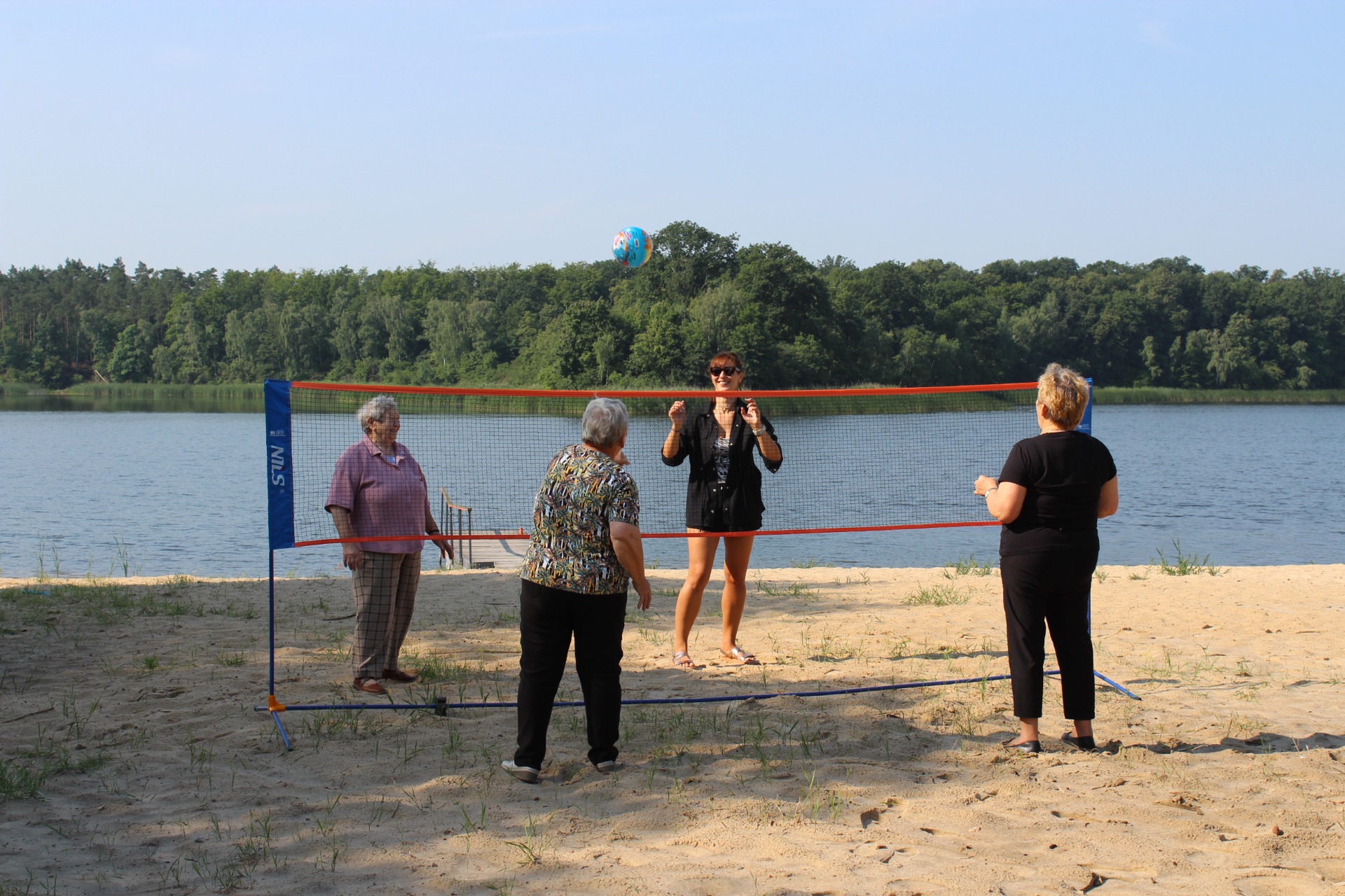 Zdjęcie wykonane nad Jeziorem Lubstowskim. Przedstawia cztery kobiety grające w piłkę siatkową na plaży. W tle woda i las.