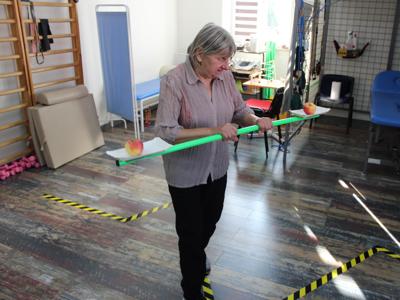 3) Zdjęcie wykonane na sali rehabilitacyjnej w DDP. Przedstawia Seniorkę pokonującą slalom tak by nie upuścić trzymanych na tackach jabłek.