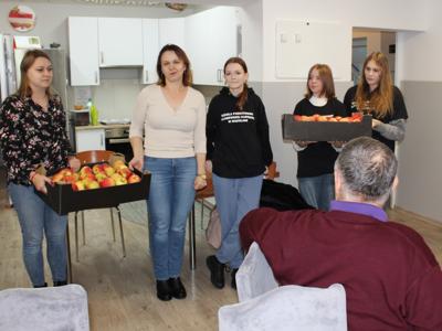 1. Zdjęcie wykonane na sali głównej Domu Dziennego Pobytu. Przedstawia Wolontariuszy wraz z opiekunami wręczających Seniorom jabłka.