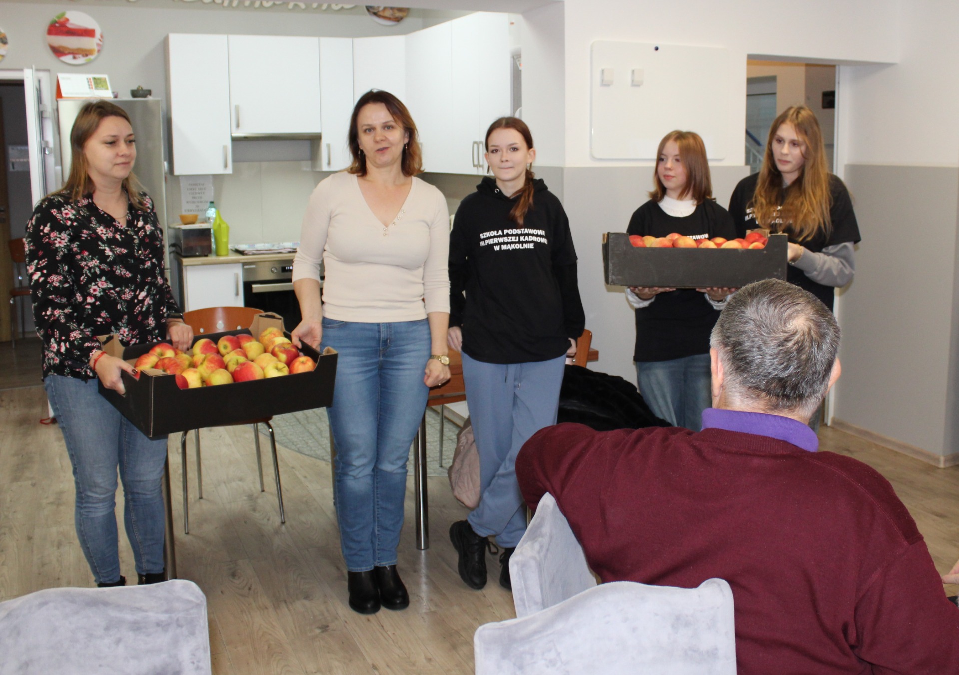 1. Zdjęcie wykonane na sali głównej Domu Dziennego Pobytu. Przedstawia Wolontariuszy wraz z opiekunami wręczających Seniorom jabłka.