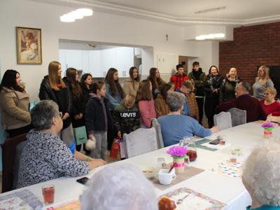 1) Zdjęcie wykonane w Domu Dziennego Pobytu. Przedstawia uczniów wraz z opiekunami, którzy przybyli do seniorów oraz pracownicę MGOPS w Sompolnie witającą uczniów.