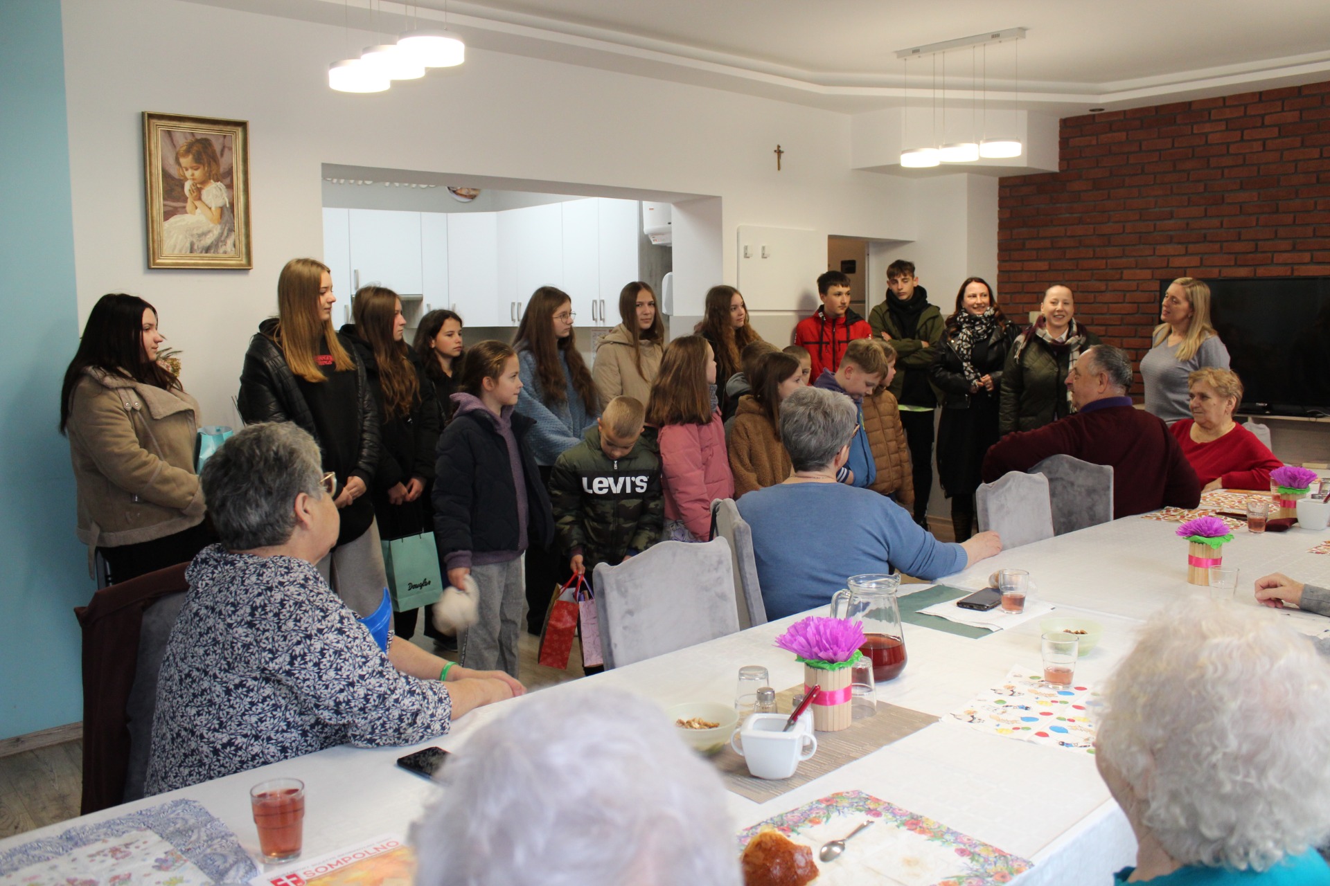 1) Zdjęcie wykonane w Domu Dziennego Pobytu. Przedstawia uczniów wraz z opiekunami, którzy przybyli do seniorów oraz pracownicę MGOPS w Sompolnie witającą uczniów.
